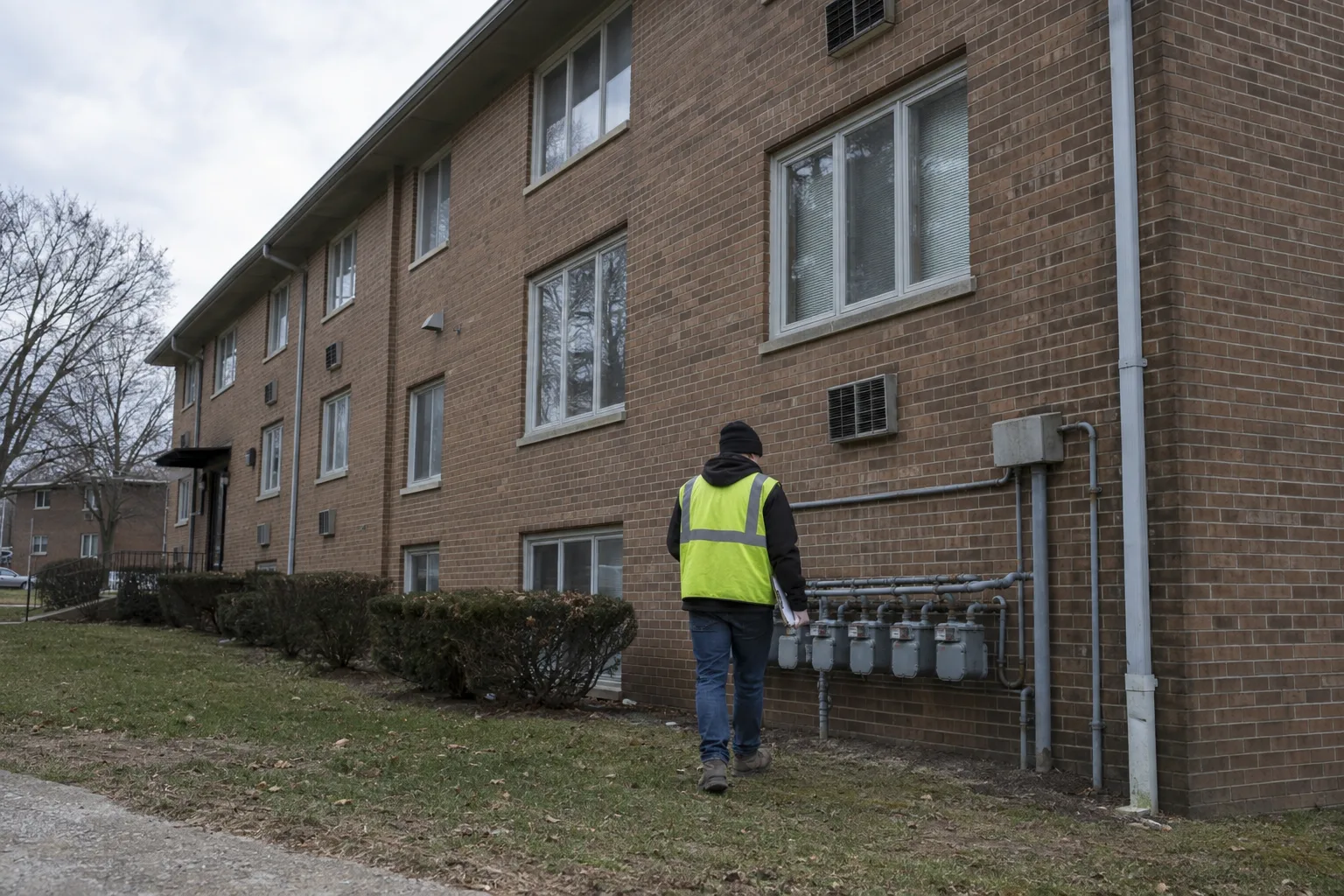 Inspector seen from behind examining the exterior façade of a brick affordable housing apartment building, holding a clipboard, under overcast afternoon light